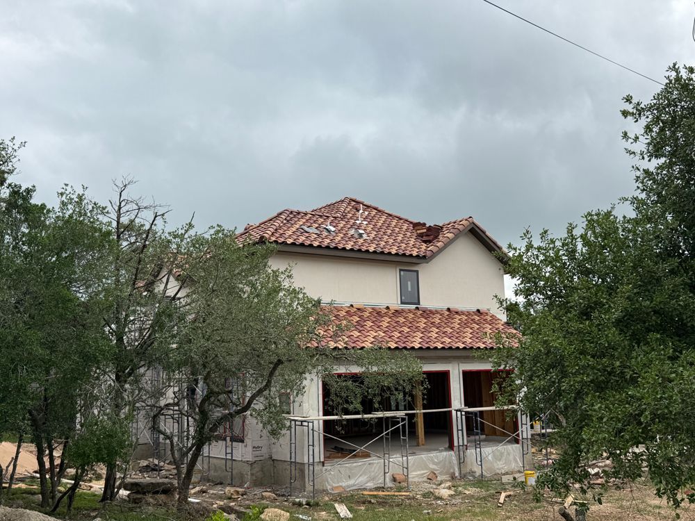 Two-story house with red tile roof and white walls, surrounded by trees under a cloudy sky