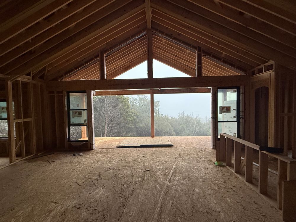 Unfinished attic room with exposed wood beams, open windows, and a view of trees outside.
