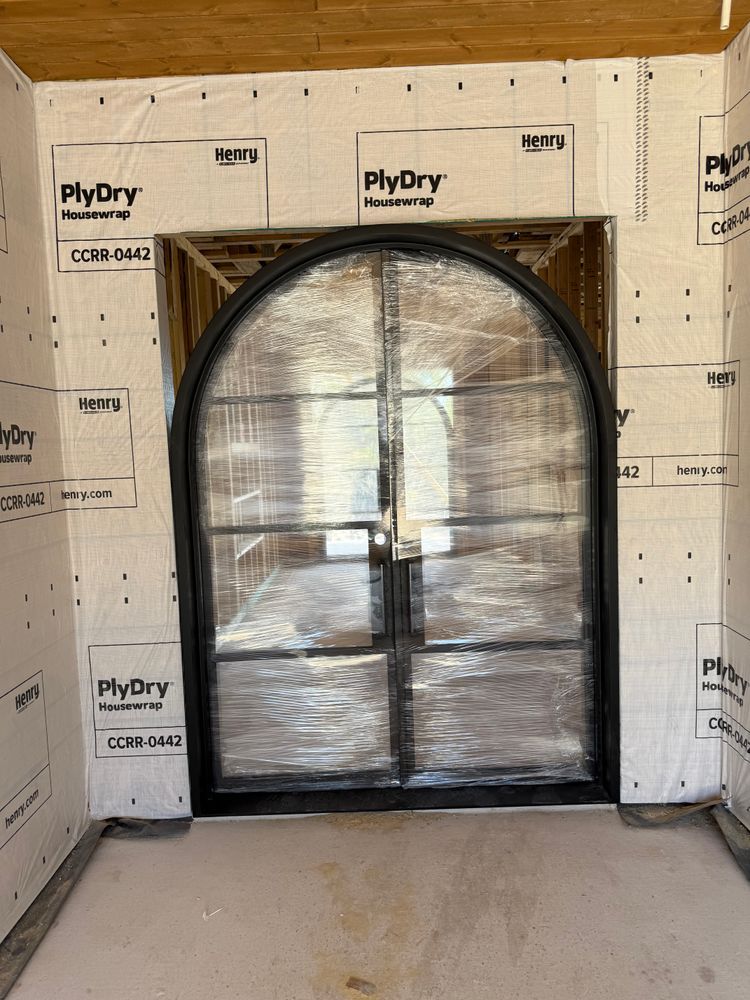 Black arched door wrapped in plastic in an unfinished room with exposed plywood walls and concrete floor