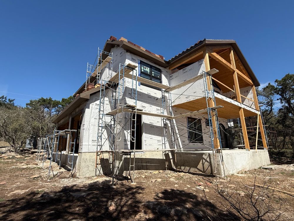 Partially built house with scaffolding under a clear blue sky, surrounded by trees and dry ground