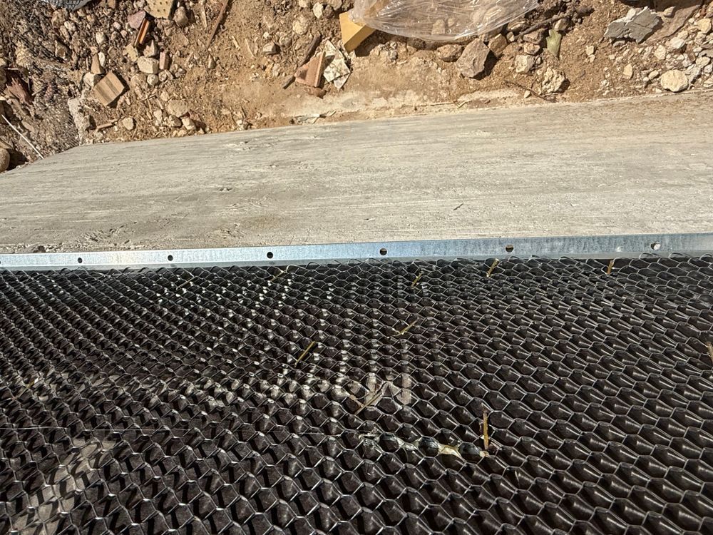 Black mesh mat beneath a concrete edge, with rocky dirt ground in the background