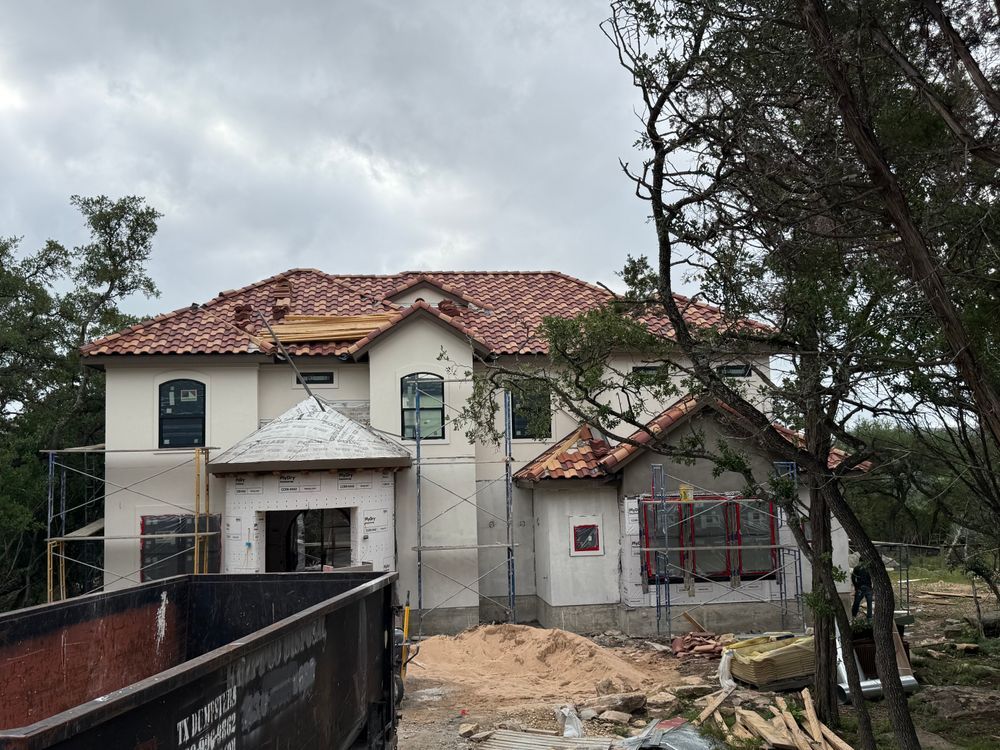 Partially built two-story house with red tile roof, scaffolding, and construction materials under cloudy skies