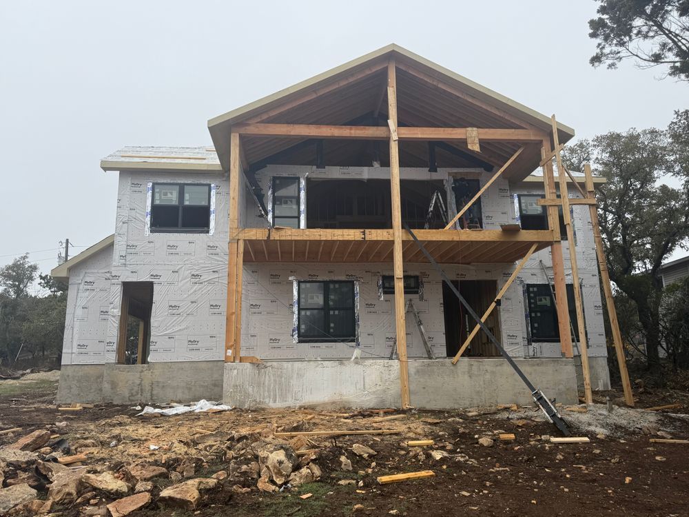 Two-story house under construction with exposed framing and stone exterior, on a dirt lot