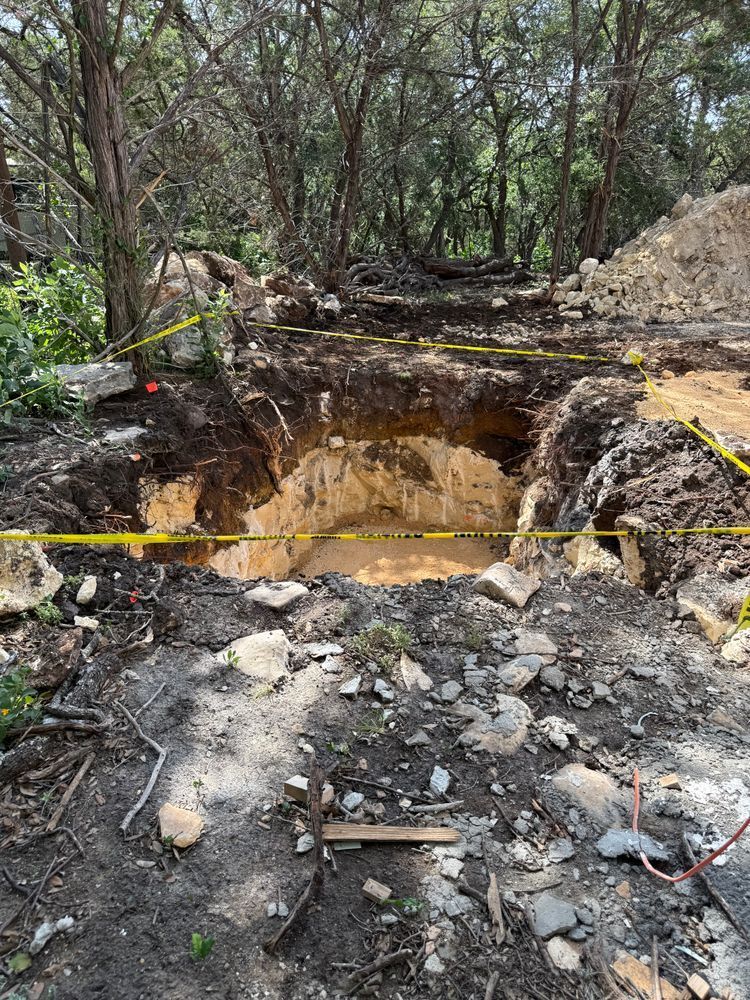 Deep dirt excavation pit in a wooded area, marked with yellow caution tape and surrounded by rubble.
