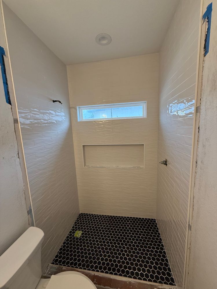 Narrow tiled shower stall with black hex floor, white textured walls, bench, and small window.