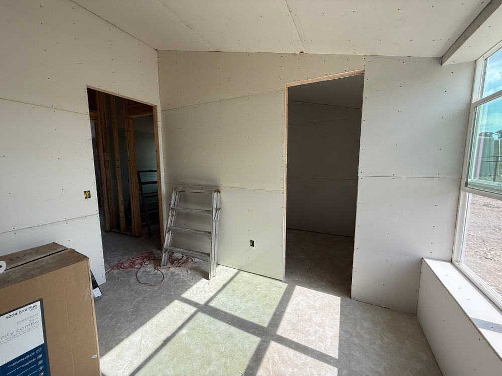 Empty room under renovation with unfinished drywall, ladder, and sunlight through large windows