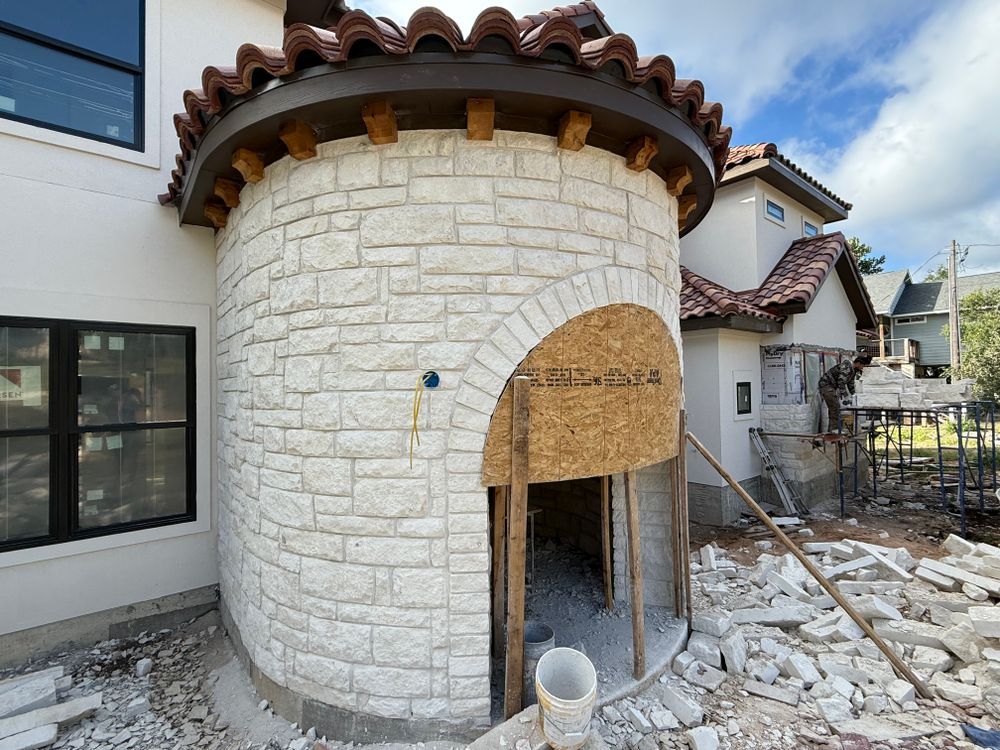 Stone exterior tower under construction with arched doorway, rubble, and scaffolding beside a house