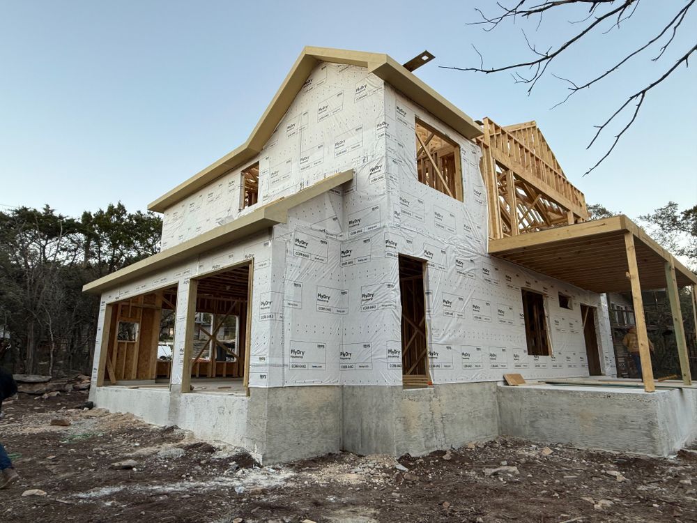 Partially built two-story house with white house wrap and exposed framing on a dirt lot