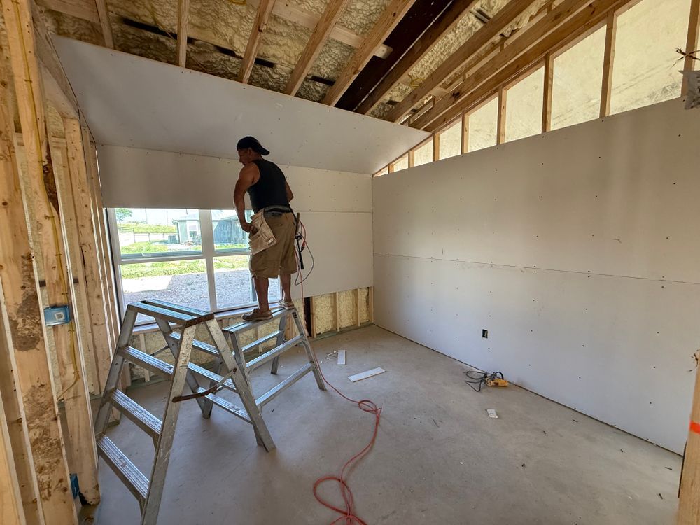 Person on a ladder installing drywall in an unfinished room with exposed framing.