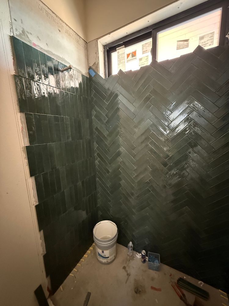 Partially tiled bathroom wall with dark green herringbone tile and a paint bucket on the floor