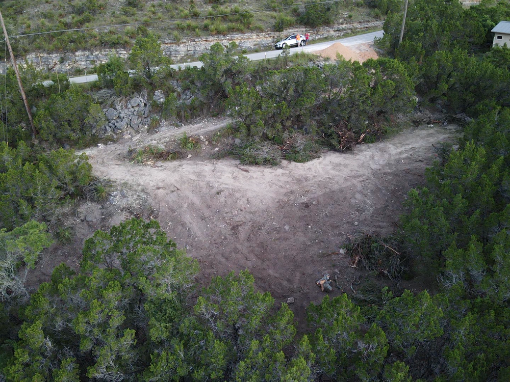 Dirt clearing in dense green brush beside a road with a small parked vehicle in the background