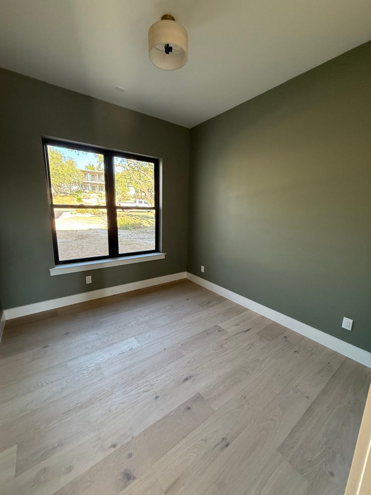 Empty small bedroom with olive green walls, light wood floor, and a window letting in daylight.