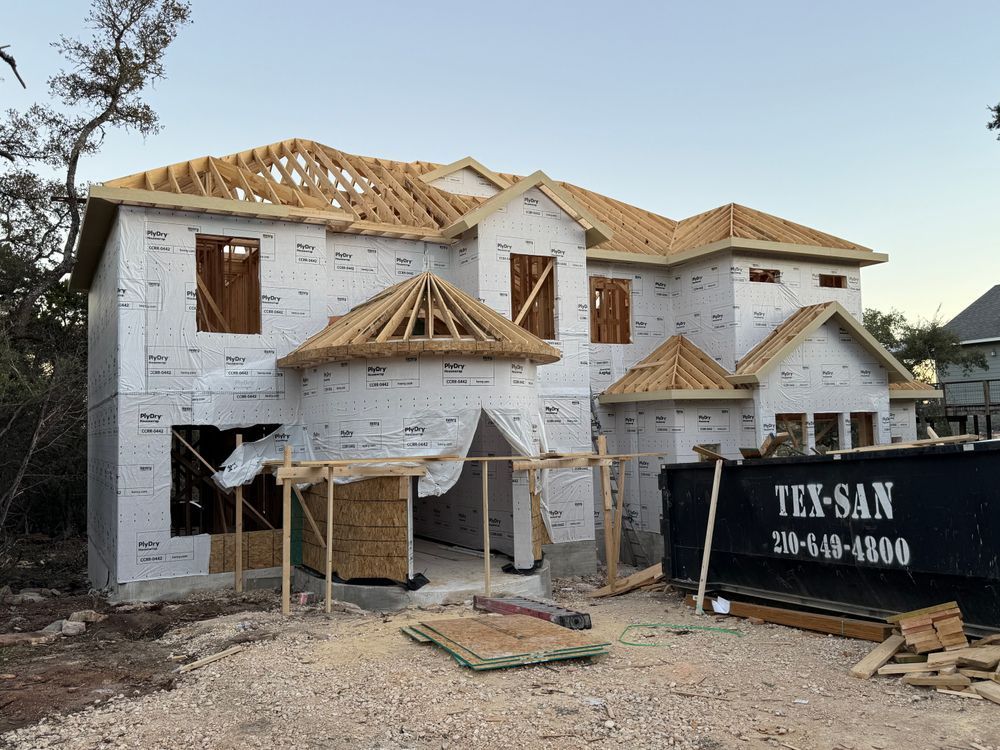Two-story house under construction with white wrap, exposed roof trusses, and a dumpster in front