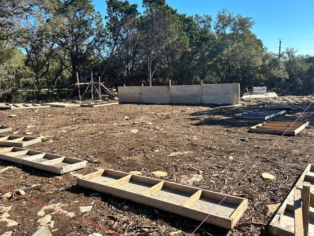 Cleared dirt lot with concrete blocks and a partially built wall under trees and blue sky