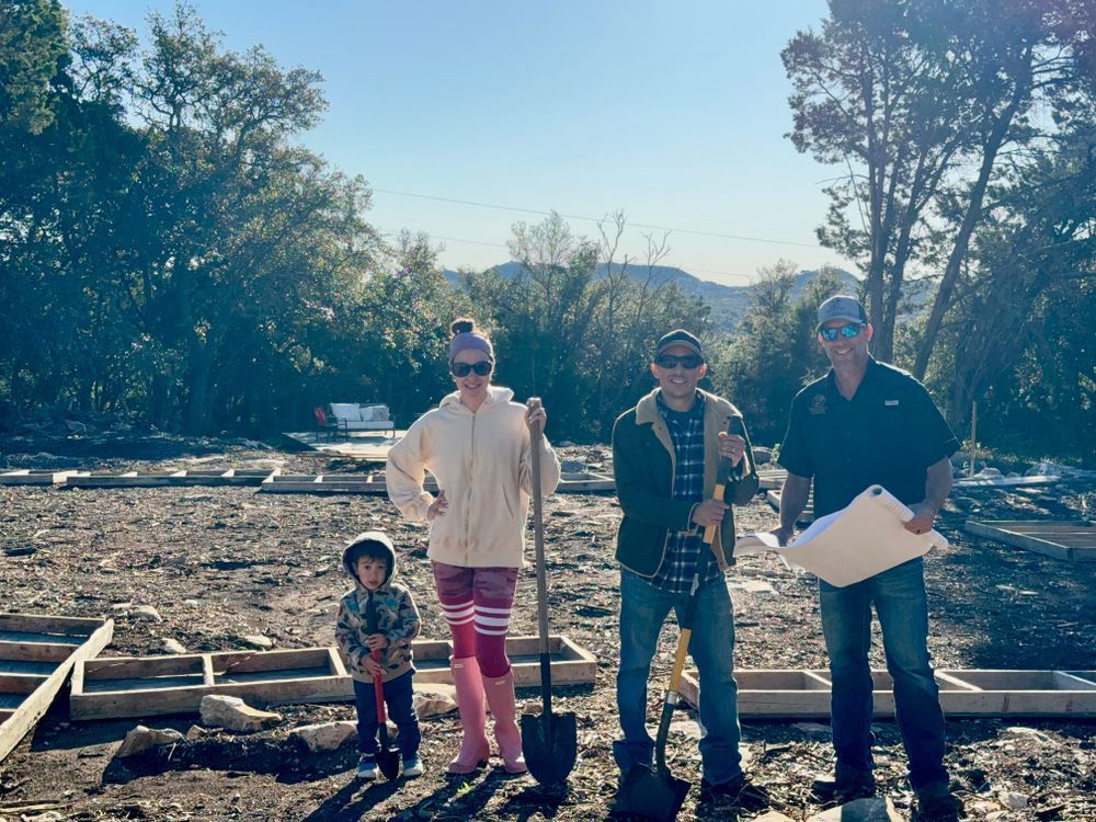 Four people standing outdoors amid debris, holding papers, with trees and a bright sky behind them.