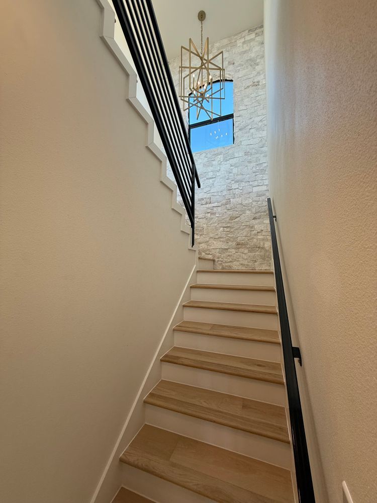Narrow indoor staircase with beige walls, black railing, and a chandelier by a blue window at the top.