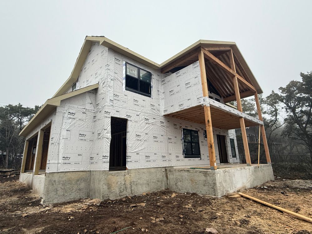 Unfinished two-story house under construction with white wrap, exposed wood roof, and concrete foundation