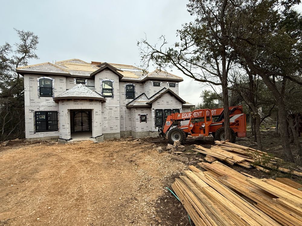 Partially built two-story house with an orange tractor and stacked lumber on a dirt lot