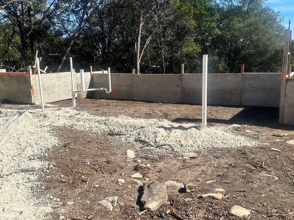 Partially built concrete foundation with upright posts in a dirt lot, surrounded by trees and a low wall.