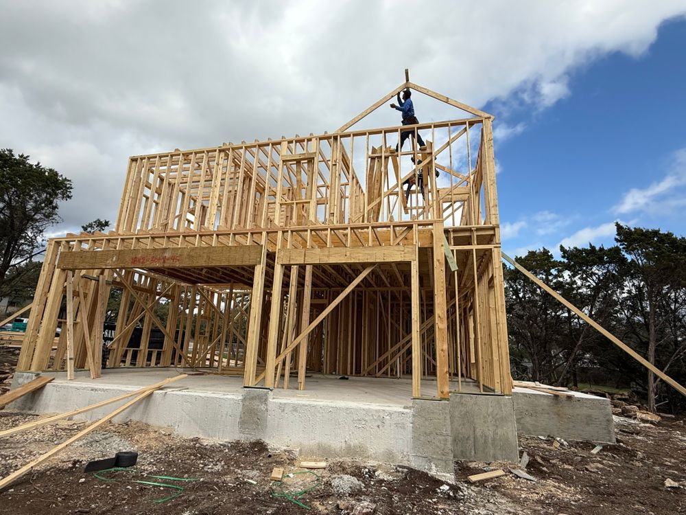 Wooden house frame under construction on a concrete foundation, with a worker on the roof against a cloudy sky