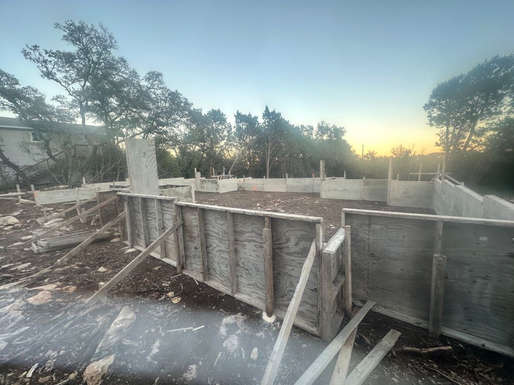 Concrete foundation forms at a construction site at sunset, with trees and a distant house in the background.