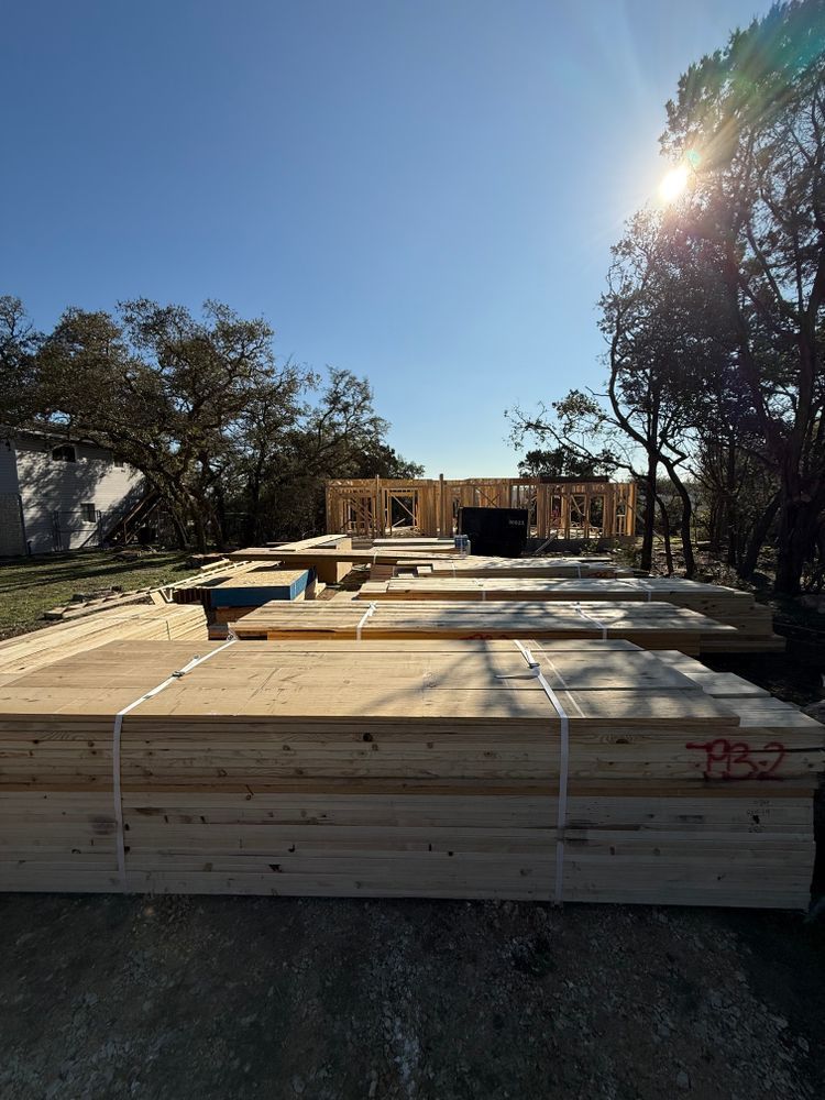 Stacked lumber on a construction site in bright sun, with framed house walls in the background.