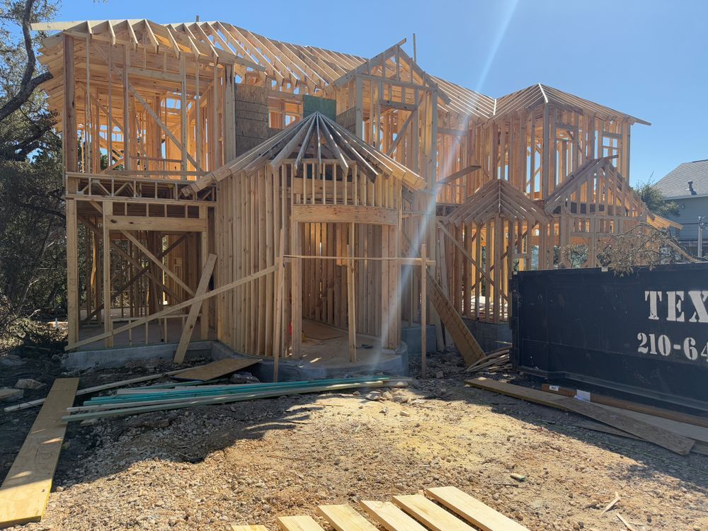 Wood-framed houses under construction on a sunny lot, with a dumpster in the foreground.