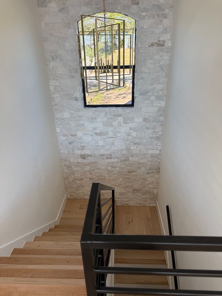 Stairwell with black handrail, white walls, and a tall window overlooking trees and a gate.