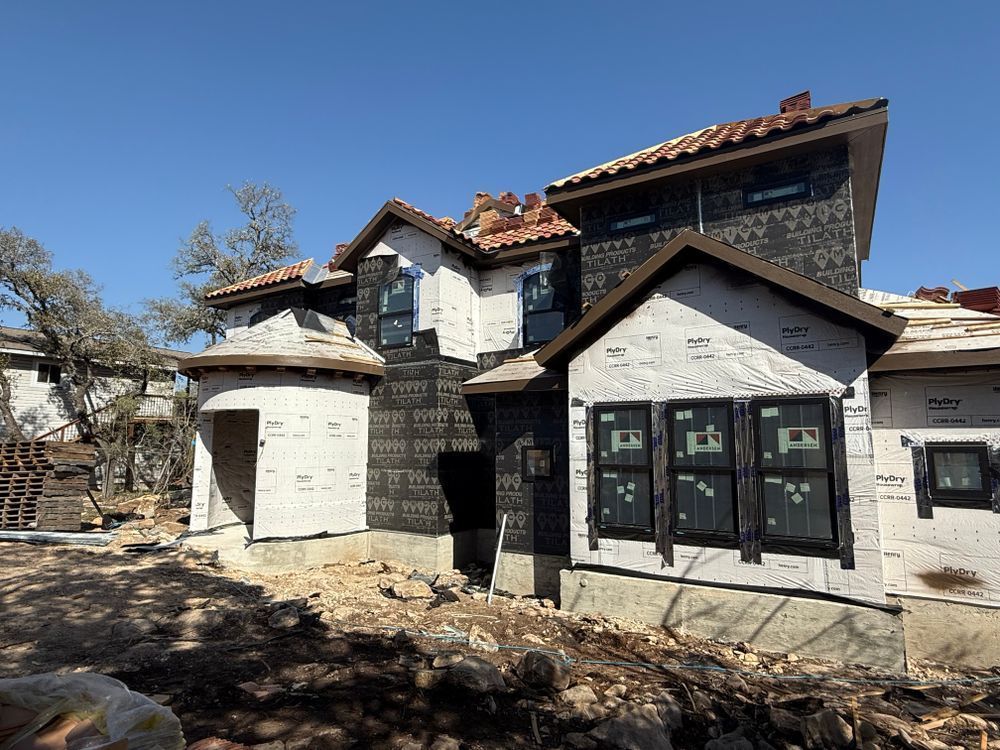 Stone house under construction with white stucco walls, dark trim, and a tiled roof in a sunny yard