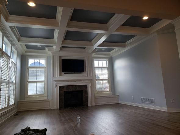 Living room with coffered ceiling, fireplace, and windows. Dark gray ceiling, white trim, wood floor, and light blue walls.
