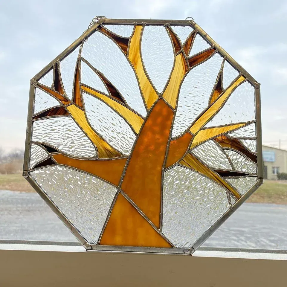 Octagonal stained glass art piece depicting a tree with amber and brown branches against a cloudy sky.