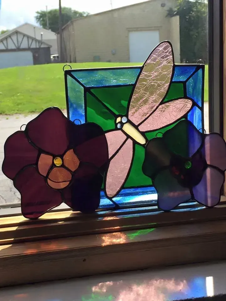 Stained glass dragonfly between two floral shapes, in a window. Colors: pink, purple, green, blue.