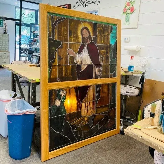 Stained glass window of Jesus, set in a wood frame, indoors. Jesus is standing, a figure kneeling below.