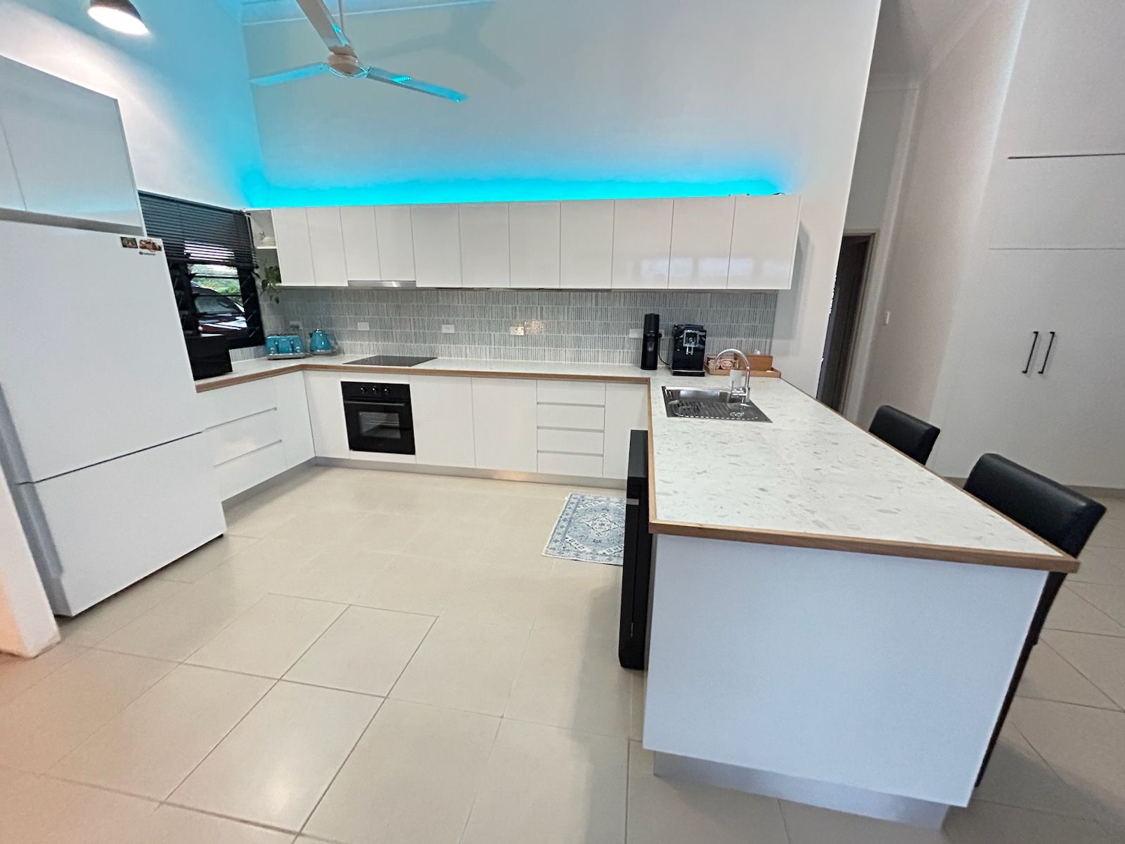 Modern white kitchen with marble-style countertops, under-cabinet blue lighting, and a breakfast bar with two stools. — Frenchy’s Plaster & Paint in Bellamack, NT