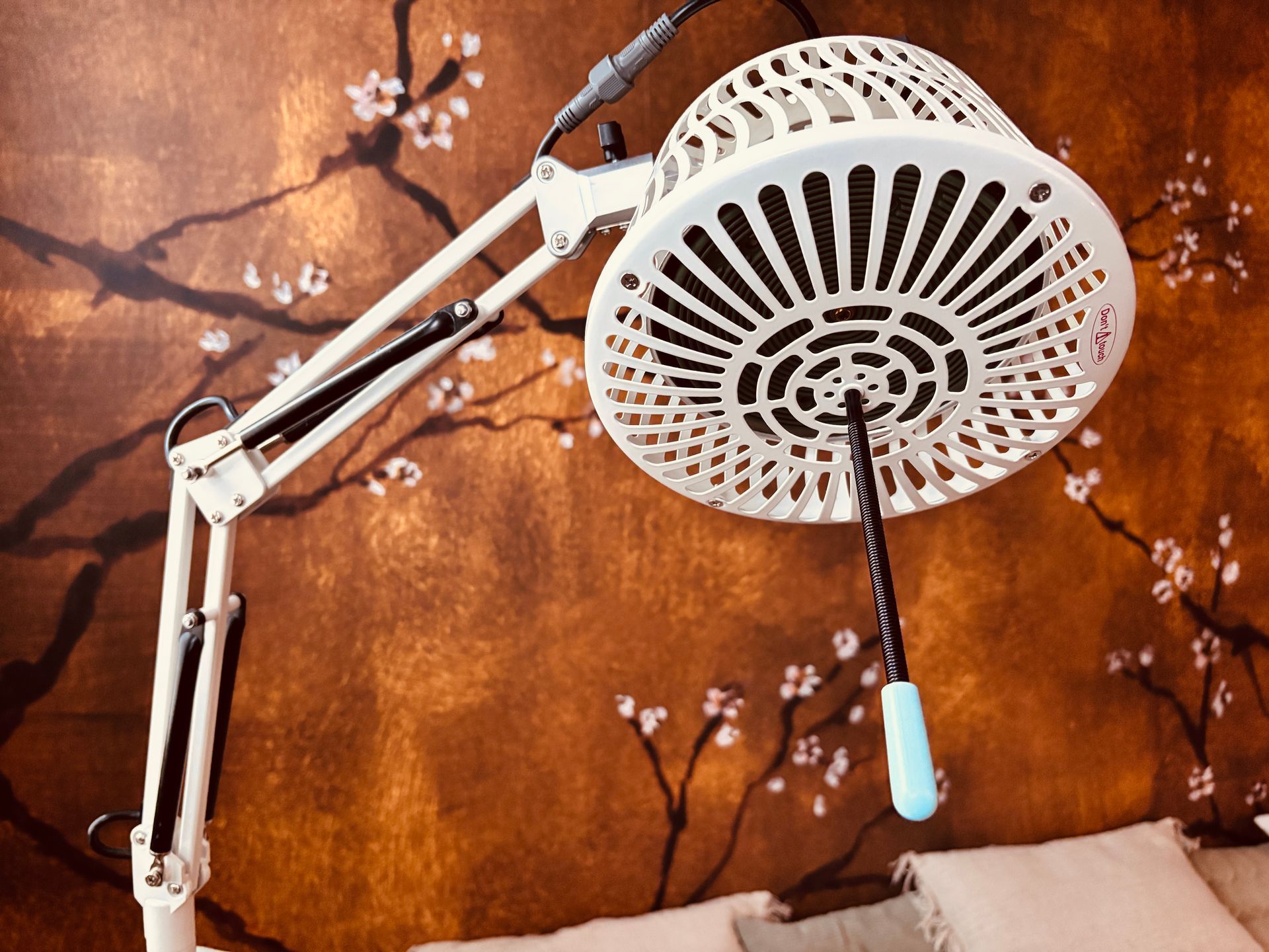 Witte, verstelbare bureaulamp met ronde, geventileerde kop tegen een oranje bloemenwand, turquoise trekkoord zichtbaar.