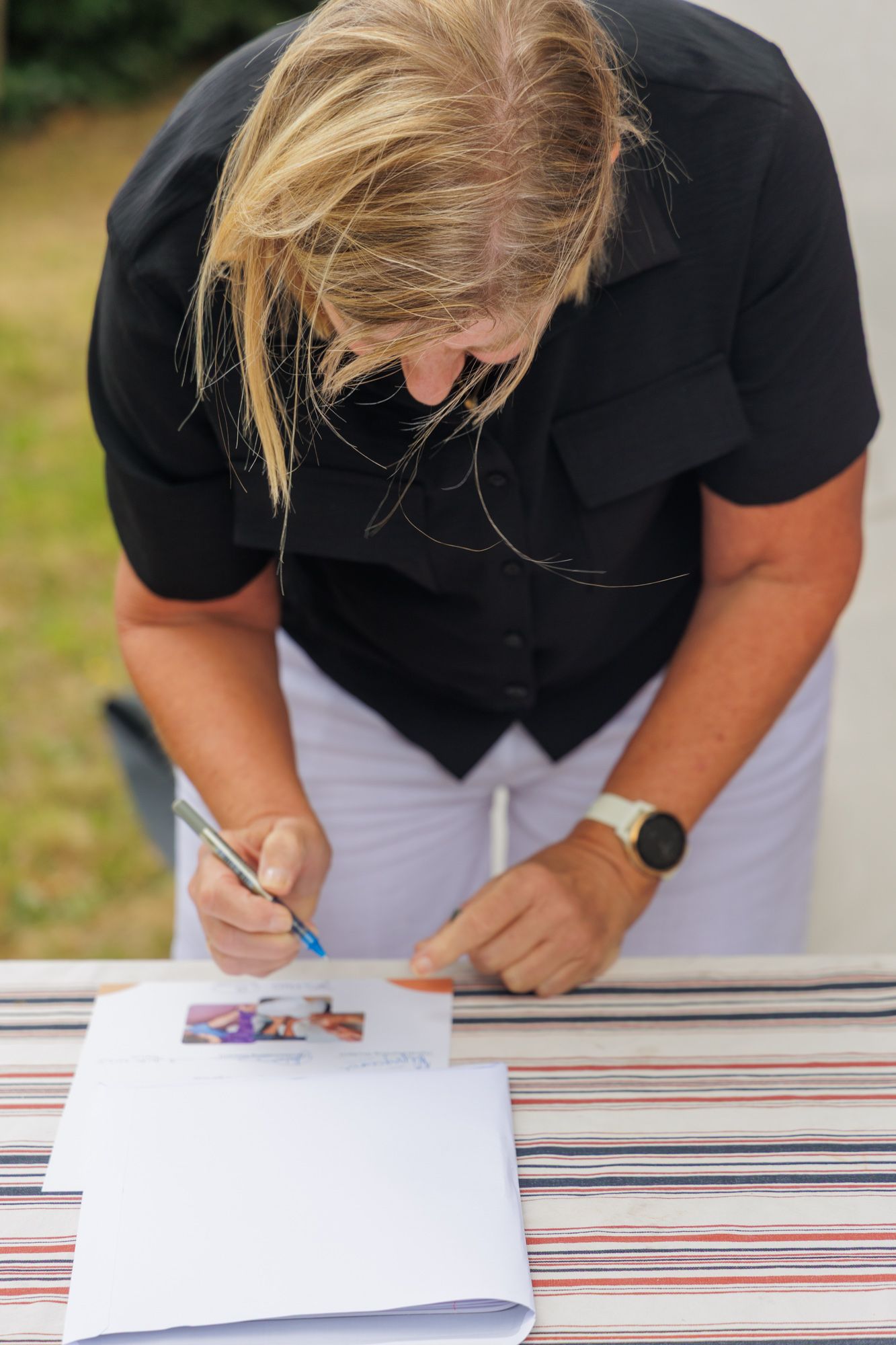 Een persoon in een zwart shirt ondertekent een document aan een tafel, voorovergebogen over witte papieren en een gestreepte tafelkleed.