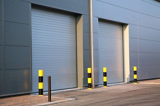 Two metal roller doors on a building, protected by black and yellow bollards on a paved surface.