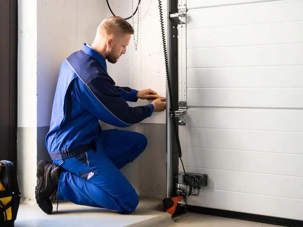 Worker in blue coveralls crouches beside a garage door, adjusting the track mechanism.