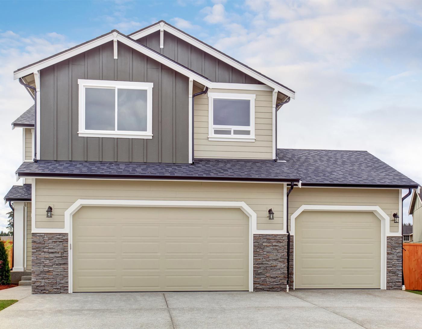 Two-story house with a tan exterior, stone accents, and two garage doors under a blue sky.