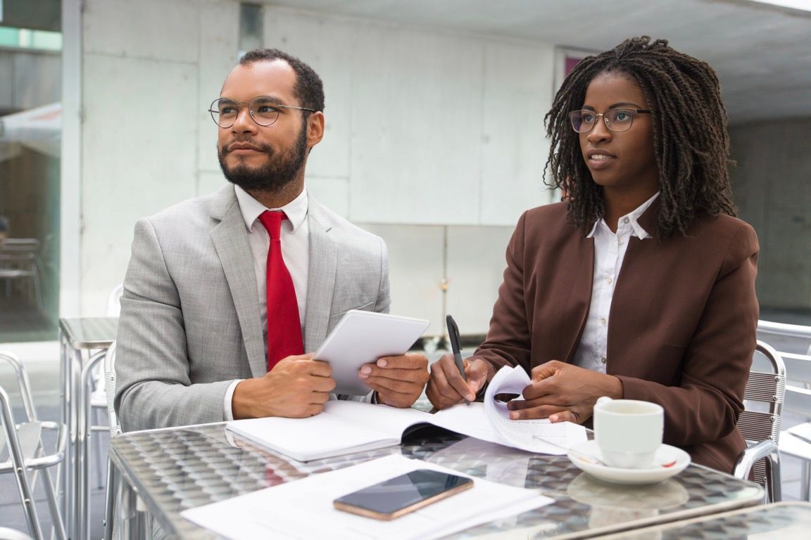 Two professionals review documents at a glass table with a laptop, smartphone, and coffee cups.