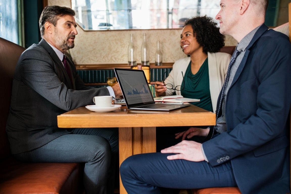 Three people in business attire discuss around a laptop at a café table
