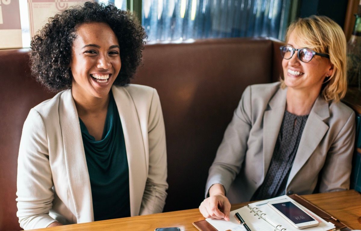 Two coworkers smiling at a table with papers and a tablet in a meeting room