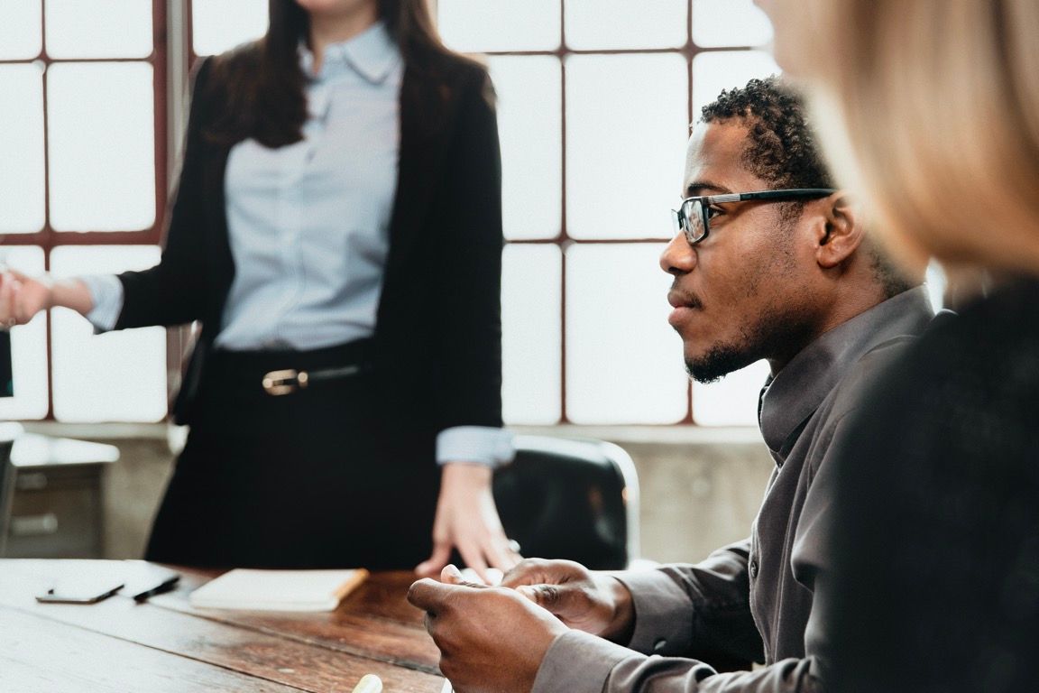 Colleagues in a meeting room, one seated man taking notes while others stand near a table.