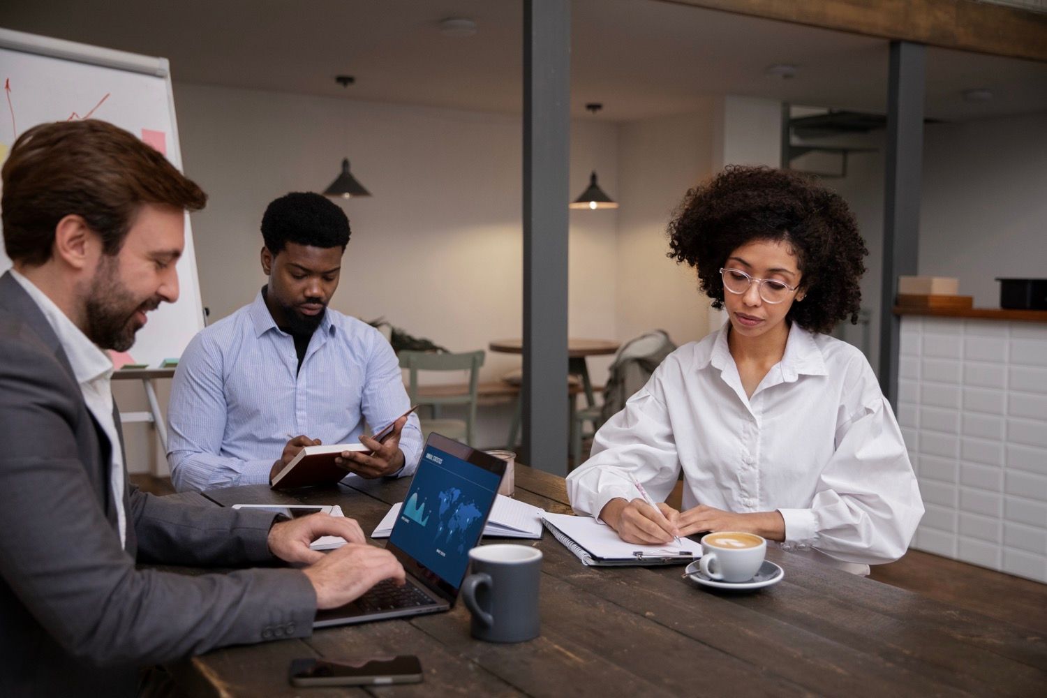Three coworkers in an office meeting, two using laptops and one taking notes at a table.