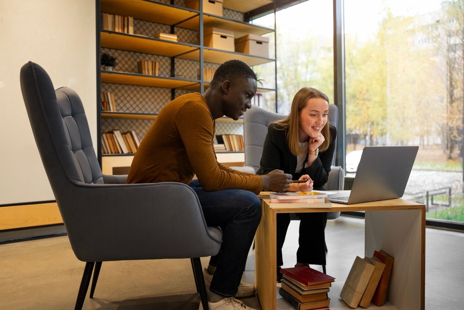 Two people working together at a laptop in a bright modern lounge with shelves and large windows