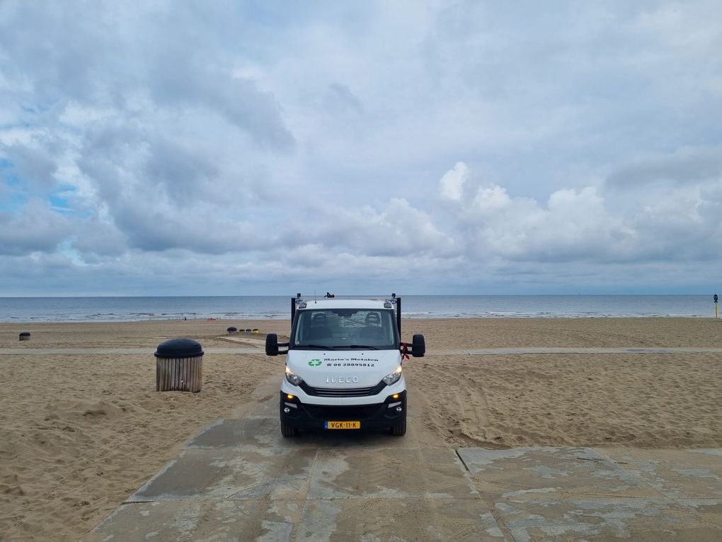 Een witte bestelbus staat geparkeerd op een zandstrand naast een betonnen pad onder een bewolkte hemel.