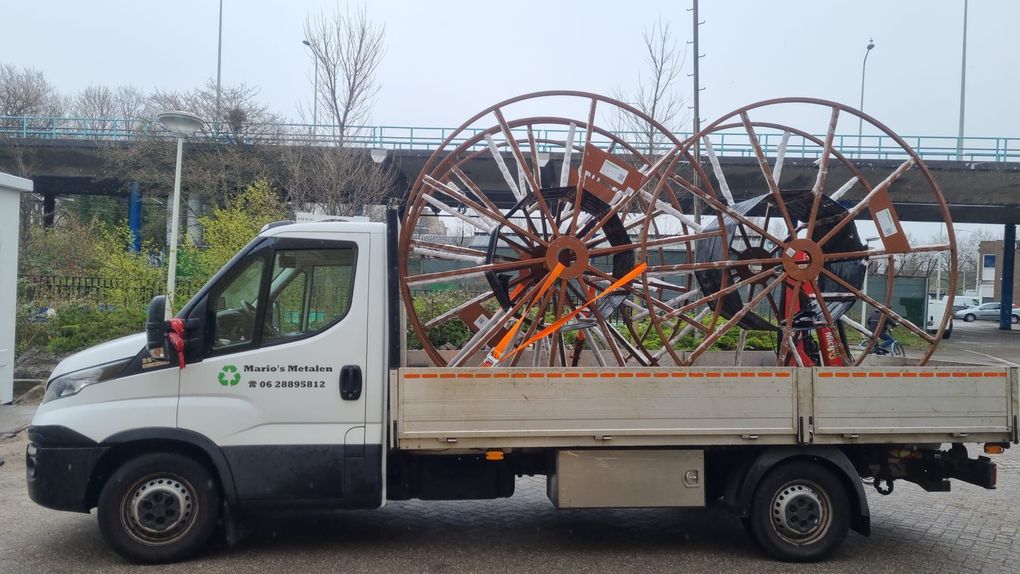 Witte vrachtwagen met open laadbak, die grote houten wielconstructies vervoert op een grindterrein buiten.