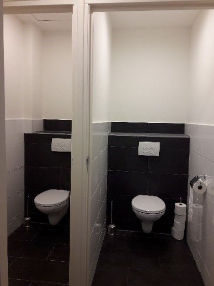 Twee kleine toiletcabines met witte toiletten, zwarte tegelwanden en toiletpapierhouders.