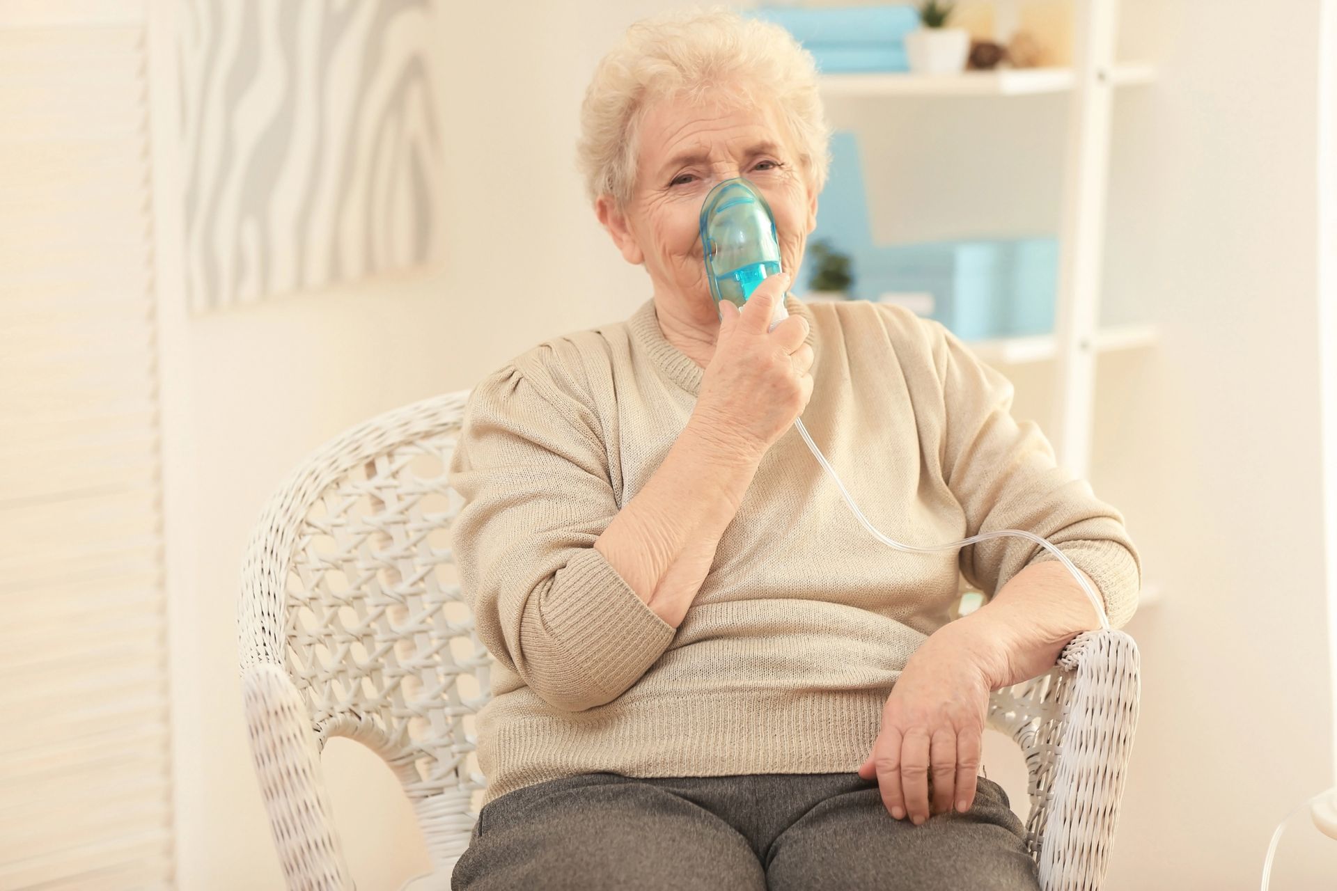 Mujer sentada en una silla de mimbre usando un inhalador en una habitación luminosa.