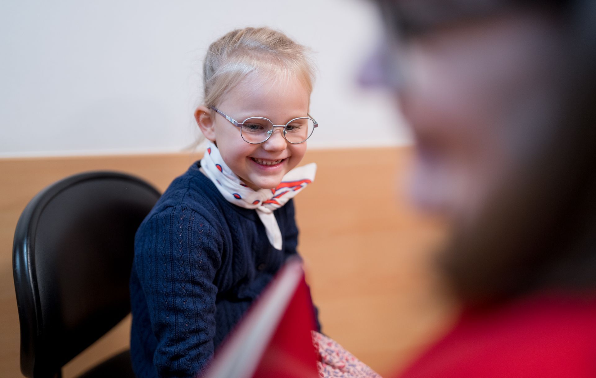 Smiling child in glasses and scarf, looking at a book held by an unseen person.